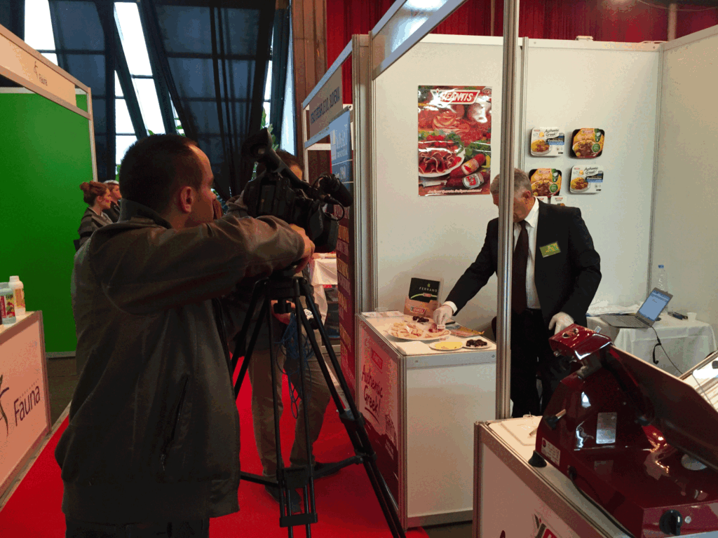 Media recording a product tasting at the Greek booth during Agrokos 2015 in Prishtina, with a representative preparing samples for presentation.