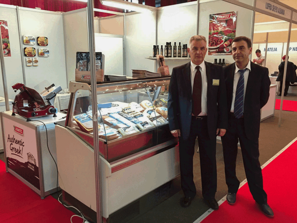 Greek food products showcased at the Agrokos 2015 exhibition in Prishtina, with company representatives standing in front of the display booth.